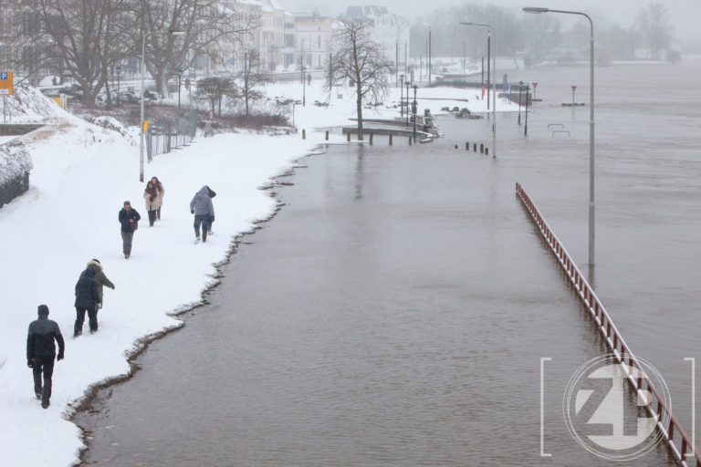 Winter in Zutphen. Mensen lopen door de sneeuw langs de hoog staande IJssel.